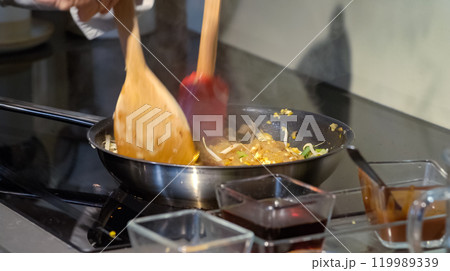 A cook stirs fry Pad Thai noodles in a pan with spatulas on electric stoves. Sauce dishes for cooking traditional Pad Thai. 119989339