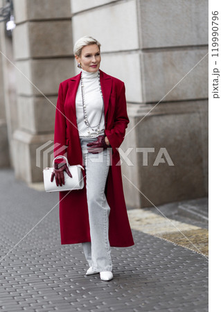Stylish Woman in Burgundy Coat Walks City Street. Wearing Fashionable clothing White Turtleneck Sweater, Pearl Necklace and Grey Jeans, Carrying White Bag. Confident and Chic Mature Fashion Model Stylish Woman in Burgundy Coat Walks City Street. Wearing Fashionable clothing White Turtleneck Sweater, Pearl Necklace and Grey Jeans, Carrying White Bag. Confident and Chic Mature Fashion Model 119990796