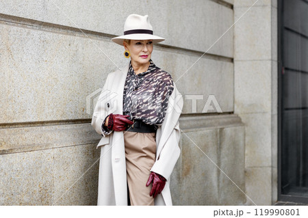 Stylish Mature Woman Wearing Fashionable Clothing Beige Coat, blouse with print, Felt Hat, Accessories and Leather Gloves Standing Against Urban Building in City, Street Style and Fashion Trends 119990801