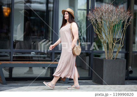 Fashionable Plus size woman walks confidently outdoors in urban city, wearing beige dress, wide brimmed hat, woven handbag. Model with long brown hair 119990820