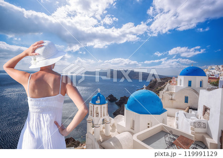 Beautiful young woman enjoying the view of Oia village with churches on Santorini island in Greece 119991129