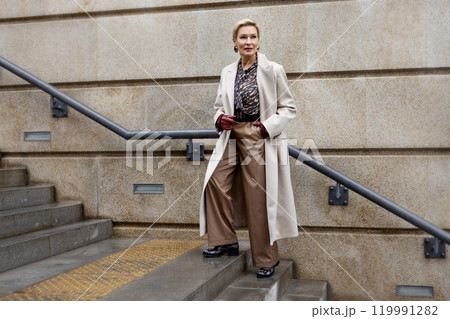 Fashionable Mature Woman Wearing Stylish Clothing Beige Coat and trousers, Accessories and Leather Gloves Standing Outdoors in City, Street Style and Fashion Trends, Winter or Autumn Outfit 119991282