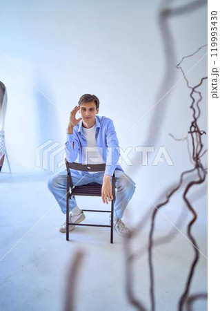 portrait of a handsome young adult man in a blue shirt sitting on a chair on a blue background portrait of a handsome young adult man in a blue shirt sitting on a chair on a blue background 119993430