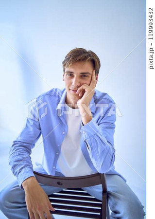 portrait of a handsome young adult man in a blue shirt sitting on a chair on a blue background 119993432