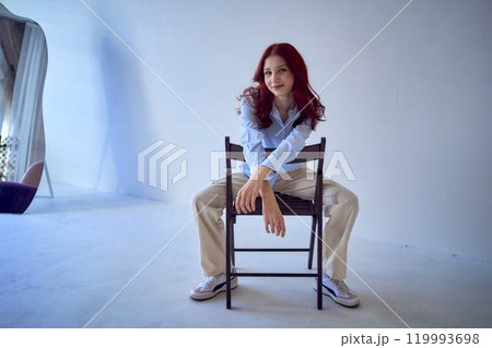 portrait of a beautiful teenage girl with red hair sitting on a chair on a white background 119993698