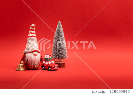 Gnome, Christmas elf in hat standing with golden bell, small car with gifts and winter tree Gnome, Christmas elf in hat standing with golden bell, small car with gifts and winter tree 119993940