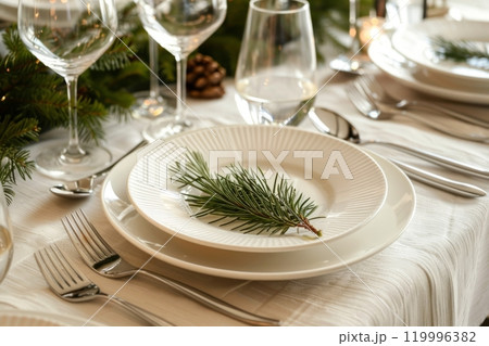 A modern Christmas dinner table setting features minimalistic decor with elegant white plates, silver cutlery, and fresh greenery accents, creating a chic and refined holiday atmosphere 119996382