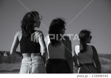 Black and white photo. Three young attractive women stand with their backs turned and their heads turned in profile Black and white photo. Three young attractive women stand with their backs turned and their heads turned in profile 119996592