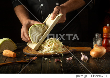 With careful precision, a chef shreds fresh cabbage on a wooden cutting board, surrounded by garlic, spices, and vibrant vegetables, creating the perfect ingredients for a delicious dish 119996784
