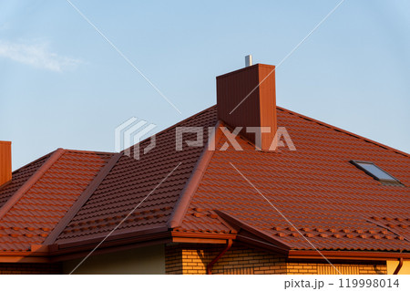 The vibrant red roof structure features a prominent chimney and an angled design, bathed in warm afternoon light against a clear blue sky, highlighting its architectural details 119998014