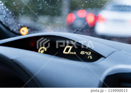 A car's dashboard features a speedometer reading zero while displaying the current time and temperature. Rain droplets are evident on the windshield, indicating wet weather 119998015