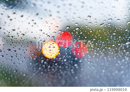 Water droplets steadily accumulate on a car window, distorting the view of vibrant traffic lights and a blurred cityscape on a gloomy, rainy day 119998019
