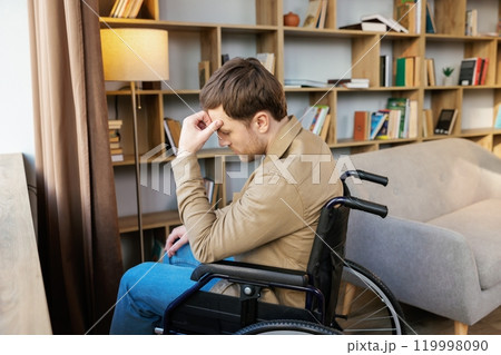 A young handsome man in a wheelchair looks out the window with a sad look A young handsome man in a wheelchair looks out the window with a sad look 119998090