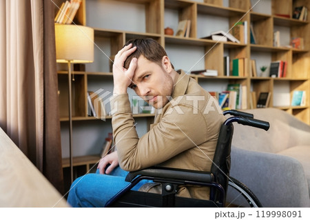 A young handsome man in a wheelchair looks out the window with a sad look A young handsome man in a wheelchair looks out the window with a sad look 119998091