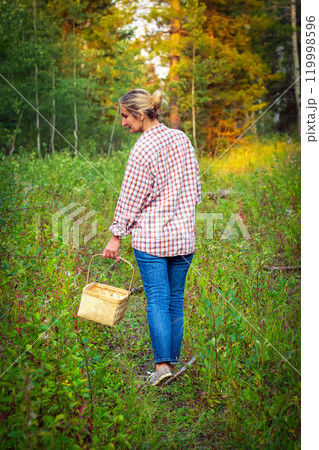 A woman in blue jeans and plaid shirt with a wicker basket is walking in the forest trail among trees and bushes and looking for mushrooms. A woman in blue jeans and plaid shirt with a wicker basket is walking in the forest trail among trees and bushes and looking for mushrooms. 119998596