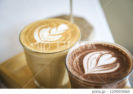 Two glasses with latte art on wooden tray are standing on white table 120003660