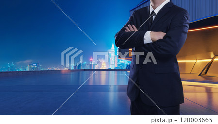 Businessman standing with arms crossed on rooftop with city skyline at night 120003665