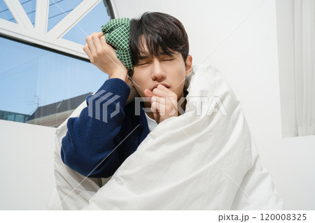 A person wrapped in a blanket and holding an ice pack on his forehead due to body aches 120008325