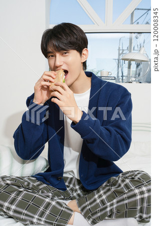 A man in his 20s sitting on the bed and eating bread A man in his 20s sitting on the bed and eating bread 120008345