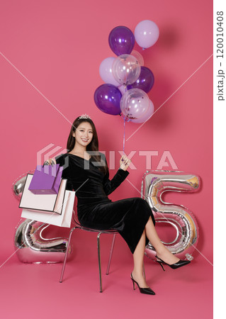 A woman in her 20s sitting and posing with a balloon and a shopping bag in front of a 25 number balloon. A woman in her 20s sitting and posing with a balloon and a shopping bag in front of a 25 number balloon. 120010408