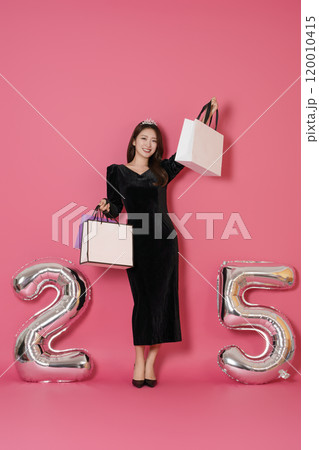 A woman in her 20s posing while holding a shopping bag in front of a 25 number balloon. 120010415