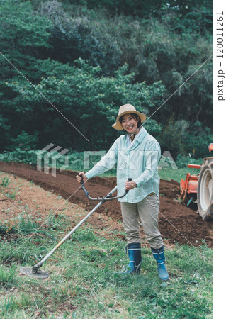 畑周りの雑草を刈払機で刈る女性 120011261