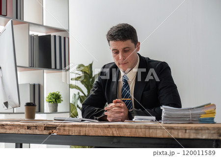 Young Businessman Deep in Thought at Modern Office Desk with Documents and Computer, Focused on Serious Work 120011589