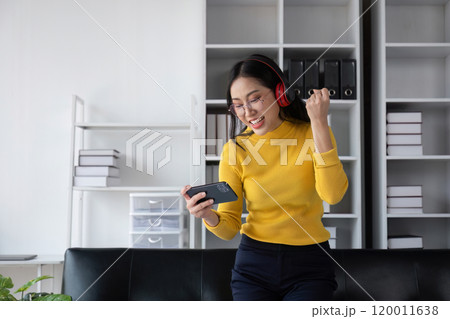 Beautiful Girl Celebrating with Joy, Wearing Headphones and Smiling While Holding a Smartphone in a Modern Office Setting 120011638