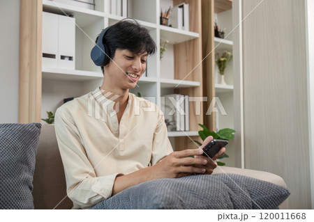 Young Man Relaxing in Modern Room While Playing on Phone and Wearing Headphones 120011668