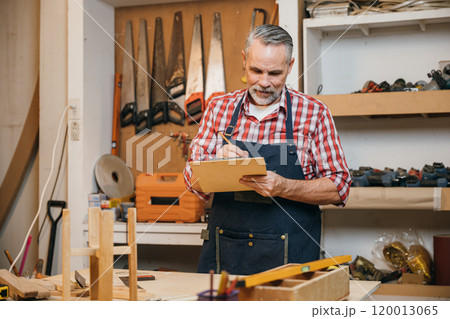 Man in plaid shirt and overalls working on a wooden project in his workshop, drawing on clipboard with pencil. Surrounded by tools and wood, woodworking and DIY craftsmanship, National Carpenters Day Man in plaid shirt and overalls working on a wooden project in his workshop, drawing on clipboard with pencil. Surrounded by tools and wood, woodworking and DIY craftsmanship, National Carpenters Day 120013065