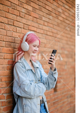 A girl with pink hair is standing next to a brick wall, holding a cell phone 120013842
