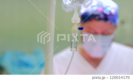 Surgeon in scrubs and mask is blurred in the background as an iv drip takes center stage, capturing the urgency of a busy hospital operating room scene Surgeon in scrubs and mask is blurred in the background as an iv drip takes center stage, capturing the urgency of a busy hospital operating room scene 120014170