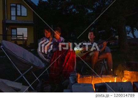 The family is sitting around the campfire, man plays the guitar, a woman listens and sings along, my daughter wrapped herself in a blanket. Family summer evening in the courtyard of the house The family is sitting around the campfire, man plays the guitar, a woman listens and sings along, my daughter wrapped herself in a blanket. Family summer evening in the courtyard of the house 120014789