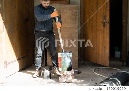 A man stirs liquid bitumen in a bucket with a wooden stick. 120019929