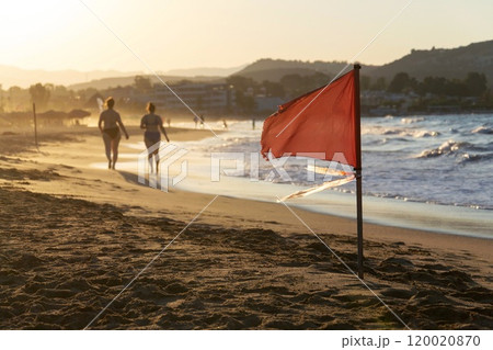 Unrecognizable people walking on a beach with a red flag warning of big waves at dawn Unrecognizable people walking on a beach with a red flag warning of big waves at dawn 120020870