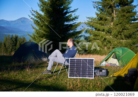 Man using photovoltaic solar panel for charging portable power station outdoor in eco-friendly camping. Male tourist sits relaxed in chair near tents. On background tall pine trees and clear blue sky. Man using photovoltaic solar panel for charging portable power station outdoor in eco-friendly camping. Male tourist sits relaxed in chair near tents. On background tall pine trees and clear blue sky. 120021069