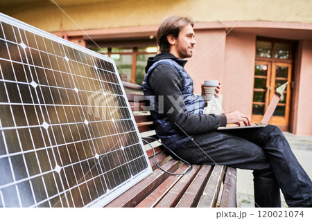 Man sits on wooden bench in urban setting, working on laptop with cup of coffee in hand. Solar panel rests beside, emphasizing sustainable, mobile workspace amidst backdrop of historic architecture. 120021074