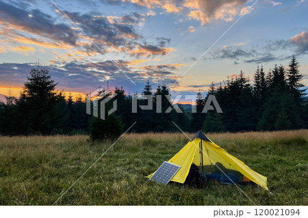 Yellow tent pitched in grassy field with solar panel beside it, capturing last light of setting sun. Backdrop of silhouetted trees and colorful sky emphasizes eco-friendly camping in nature. Yellow tent pitched in grassy field with solar panel beside it, capturing last light of setting sun. Backdrop of silhouetted trees and colorful sky emphasizes eco-friendly camping in nature. 120021094