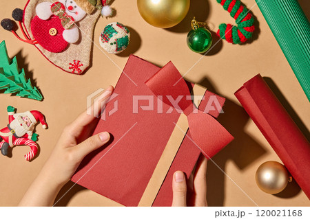 Top-down close-up photo Christmas theme showing a hand holding a red gift box. On a brown surface are Christmas themed decorations such as candy cane, Christmas balls, paper pines, and rolls of paper. Top-down close-up photo Christmas theme showing a hand holding a red gift box. On a brown surface are Christmas themed decorations such as candy cane, Christmas balls, paper pines, and rolls of paper. 120021168