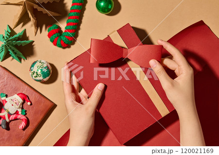 Close-up picture taken from above Christmas decorations including candy cane, noel balls, and paper pines are shown on a beige background with a hand gripping and fixing a ribbon on a red present box. Close-up picture taken from above Christmas decorations including candy cane, noel balls, and paper pines are shown on a beige background with a hand gripping and fixing a ribbon on a red present box. 120021169