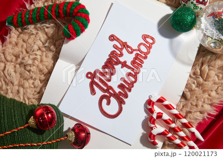 A Christmas card with cute decorative objects such as candy canes, wool scarves, Noel balls, and snow globes displayed in a fur rug on a red background, an idea for a Christmas photo template. A Christmas card with cute decorative objects such as candy canes, wool scarves, Noel balls, and snow globes displayed in a fur rug on a red background, an idea for a Christmas photo template. 120021173