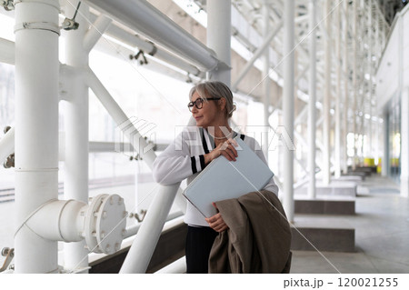 An older woman in a stylish outfit stands in a modern building, holding a laptop and a coat while An older woman in a stylish outfit stands in a modern building, holding a laptop and a coat while 120021255