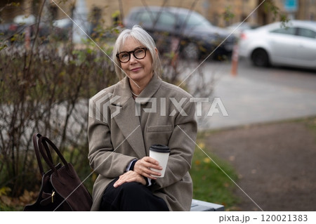 An older woman enjoys a warm drink while seated on a park bench during a cool autumn afternoon 120021383