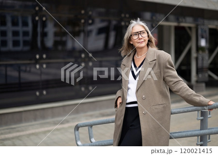 Confident older woman in stylish attire leans casually on railing outside a modern building during Confident older woman in stylish attire leans casually on railing outside a modern building during 120021415