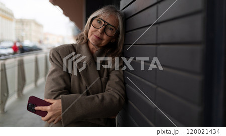 Elderly businesswoman standing outside an office, engaging with her smartphone on a cool day in the 120021434