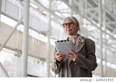 Elderly businesswoman standing confidently outside her office while holding a tablet and preparing 120021454