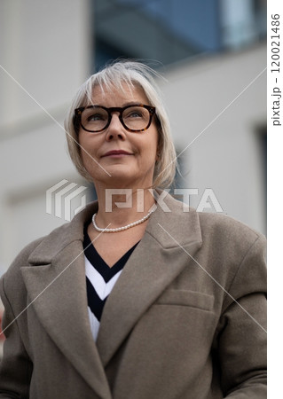 Stylish elderly woman with glasses and a chic coat standing confidently outdoors in a modern urban Stylish elderly woman with glasses and a chic coat standing confidently outdoors in a modern urban 120021486