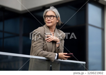 Elderly businesswoman in a stylish outfit stands on a balcony while holding her smartphone and 120021902