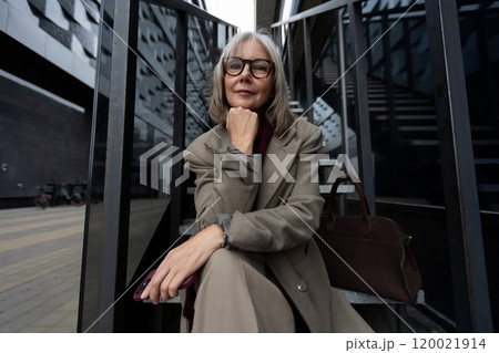 An older woman poses thoughtfully on a staircase outside a modern building in an urban setting An older woman poses thoughtfully on a staircase outside a modern building in an urban setting 120021914