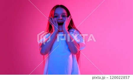 Girl with surprised expression holding hands on cheeks and looking excited in neon light against vivid gradient background. 120022857